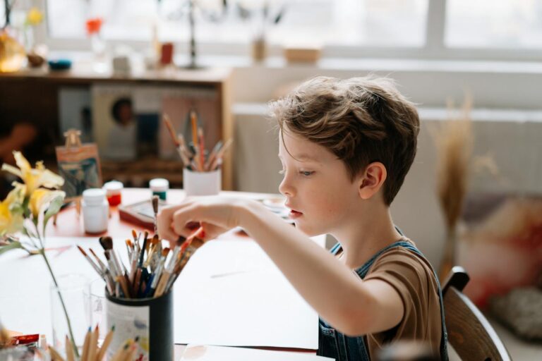 A young boy engages in painting in a sunlit art studio, surrounded by colorful brushes and supplies.